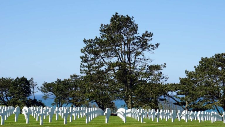 Soldatenfriedhof mit mehrere Reihen weißer Marmorkreuze, im Hintergrund Bäume und das Meer