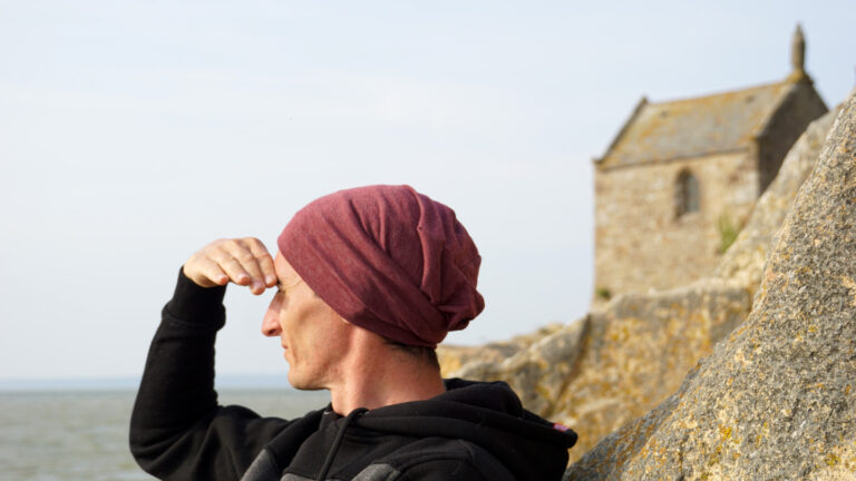 Mann mit rotem Beanie sitzt an einem Felsen vor einer Kapelle, schütz die Augen vor dem Sonnenlicht und blickt aufs Meer