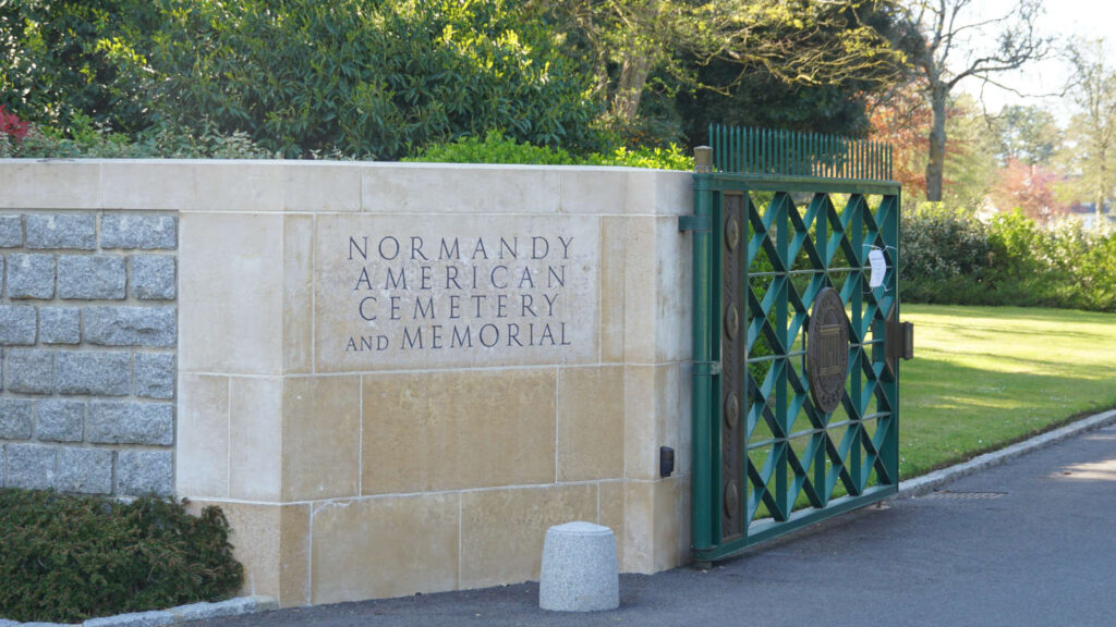 grünes Eingangstor mit der englischen Aufschrift "Normandy American Cemetery and Memorial"