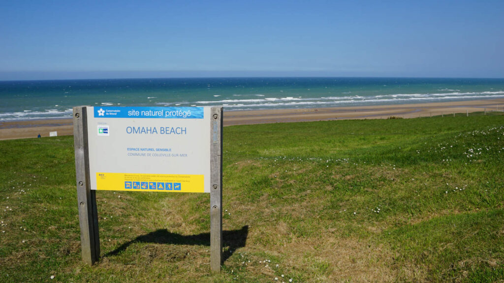 im Vordergrund eine Infotafel mit der Aufschrift Omaha Beach, im Hintergrund der Strand und das wellengepeitschte Meer