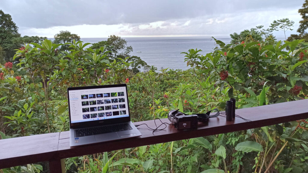 Laptop, Fotokamera und Fernglas auf einer Veranda mit Ausblick auf eine Meeresbucht