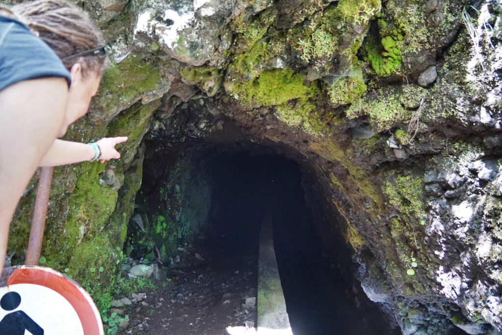 Frau zeigt in die Tunnelöffnung, wo ein winzig kleiner Lichtpunkt das Ende markiert, Levada das Rabaças auf Madeira